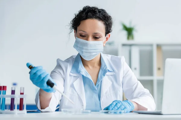 Cientista afro-americano em máscara médica segurando pipeta borrada perto da placa de Petri e laptop em primeiro plano borrado — Fotografia de Stock