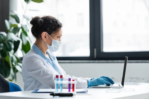 Vista lateral do cientista afro-americano em máscara médica e luvas de látex usando laptop perto de tubos de ensaio desfocados — Fotografia de Stock