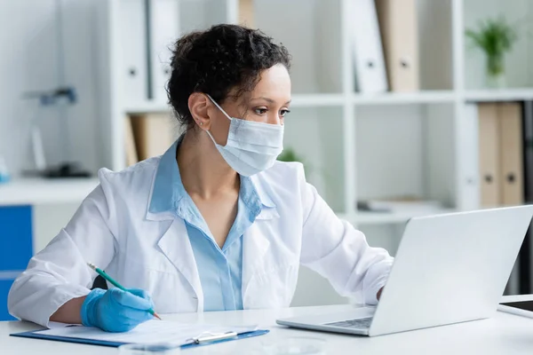 African american scientist in medical mask using laptop and writing on clipboard near blurred petri dishes — Stock Photo