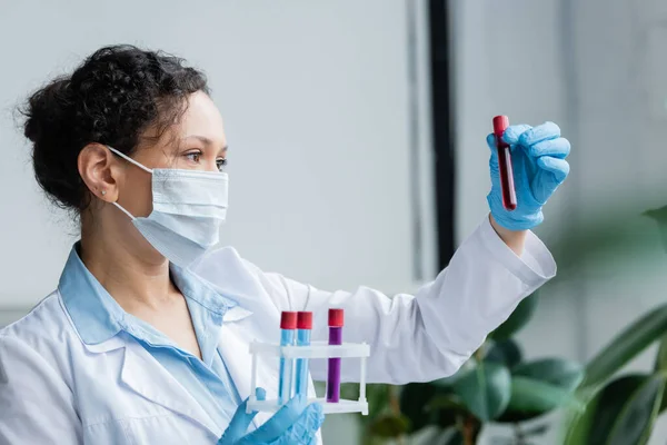 Cientista afro-americano em máscara médica olhando para tubo de ensaio com amostra — Fotografia de Stock