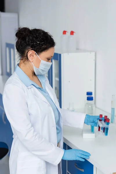 Side view of african american scientist in protective mask and gloves holding test tubes in laboratory — Stock Photo