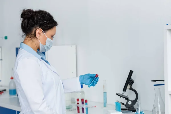 Vue latérale d'un scientifique afro-américain en masque médical et manteau blanc tenant un tube à essai près de flacons en laboratoire — Photo de stock