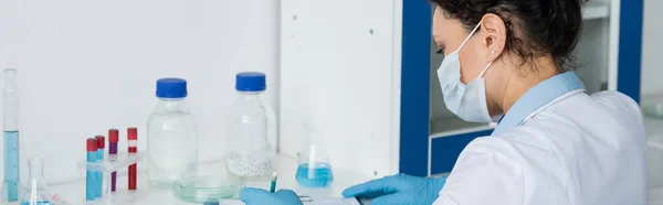 African american scientist in medical mask writing on clipboard near test tubes, banner — Stock Photo