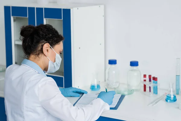 African american scientist in medical mask and latex gloves writing on clipboard near flasks and test tubes — Stock Photo