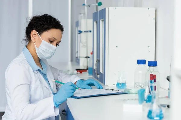 Scienziato afroamericano in maschera medica guardando appunti vicino a fiaschi e provette in laboratorio — Foto stock