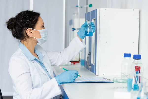 Vista lateral do cientista afro-americano em máscara médica segurando frasco perto da área de transferência — Fotografia de Stock