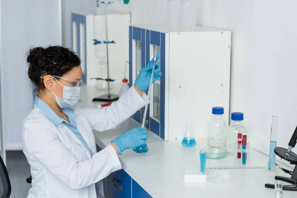 Vista lateral do cientista afro-americano em máscara médica e óculos de segurança segurando pipeta e frasco perto do microscópio — Fotografia de Stock