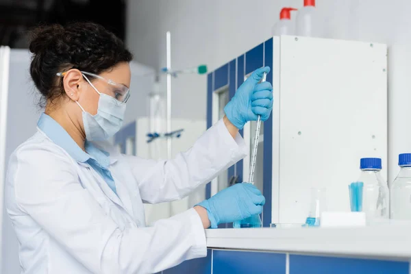 Vista lateral do cientista afro-americano em óculos de segurança segurando pipeta e frasco em laboratório — Fotografia de Stock