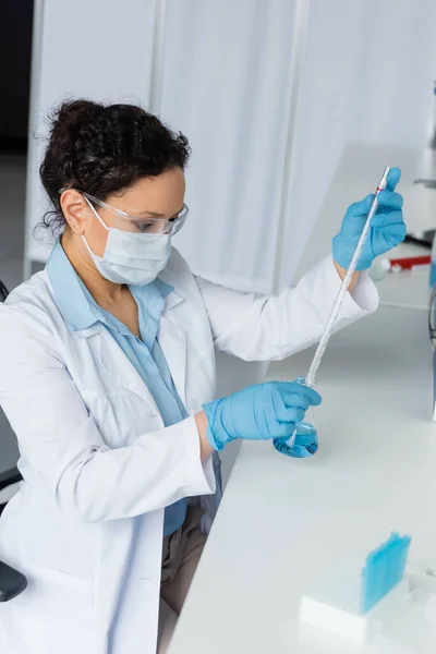 African american scientist in protective goggles holding pipette and flask near blurred test tubes - foto de stock