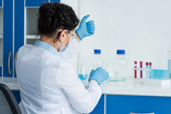 Scientifique afro-américain en masque médical et gants en latex tenant une pipette floue en laboratoire — Photo de stock