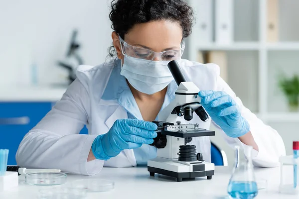 Scientifique afro-américain en masque médical et gants de latex regardant au microscope près des boîtes de Pétri — Photo de stock