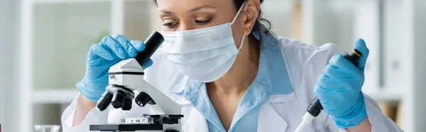 Cientista afro-americano em máscara médica segurando pipeta eletrônica perto do microscópio, banner — Fotografia de Stock