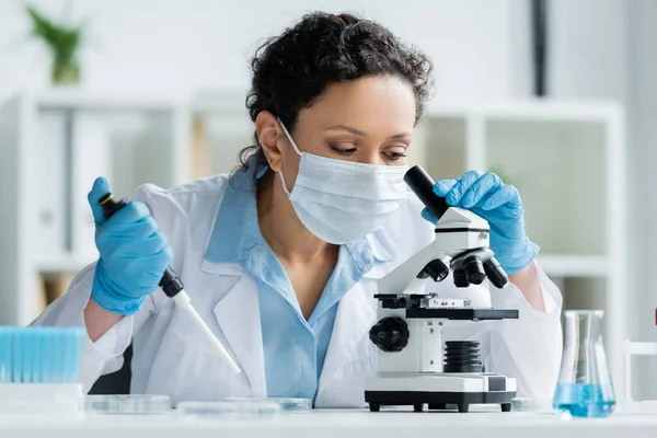 Scientifique afro-américain en masque médical tenant une pipette électronique et regardant au microscope en laboratoire — Photo de stock
