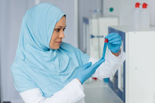 Muslim scientist in hijab and latex gloves looking at sample in test tube — Stock Photo