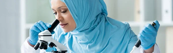 Muslim scientist in medical mask holding pipette and looking through microscope, banner — Stock Photo