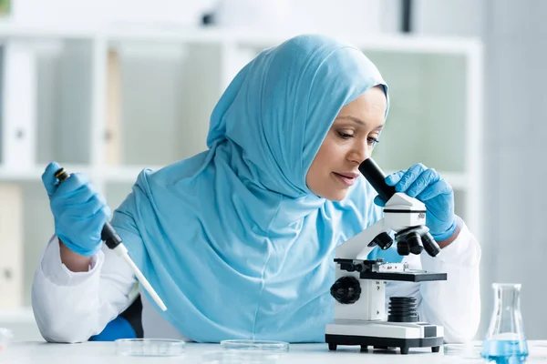 Arabian scientist in hijab looking through microscope and holding electronic pipette — Stock Photo