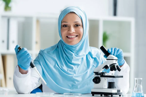 Усміхнений арабський вчений тримає піпетку і мікроскоп під час роботи в лабораторії — стокове фото