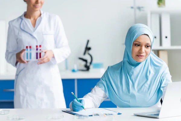 Muslim scientist writing on clipboard and looking at laptop near blurred colleague with test tubes — Stock Photo