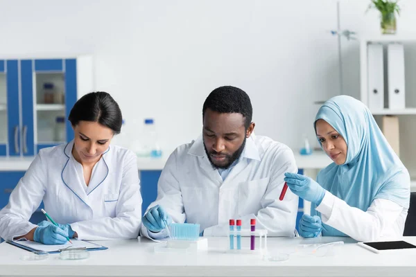Interracial scientists working with test tubes near colleague writing on clipboard in lab — Stock Photo