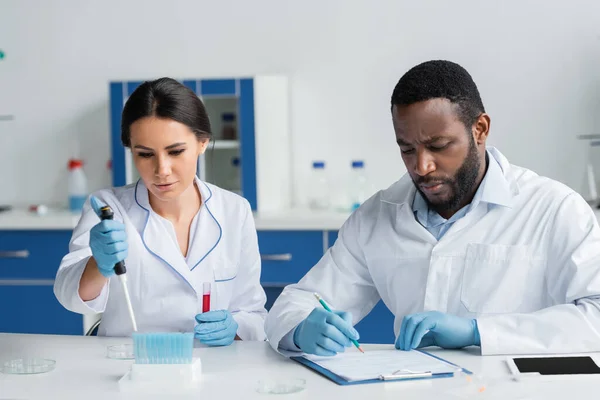 African american scientist writing on clipboard near colleague with pipette and test tubes — Stock Photo