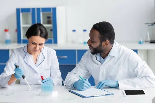 Smiling african american scientist in latex gloves writing on clipboard and looking at colleague with test tube - foto de stock