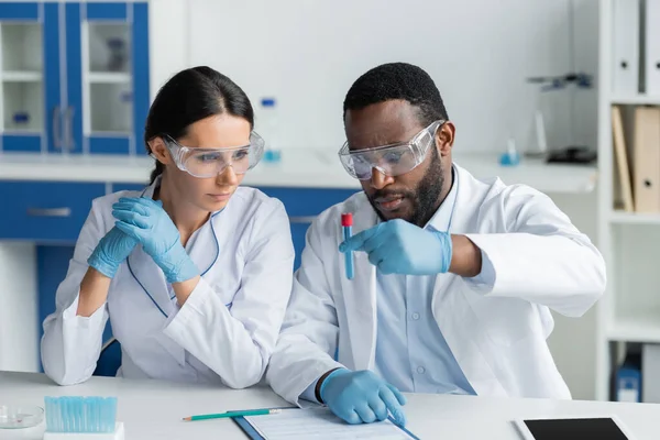 Interracial scientists in safety goggles holding test tube near clipboard and digital tablet — Stock Photo