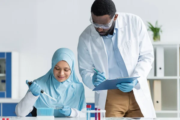 African american scientist in safety goggles writing on clipboard near smiling arabian colleague with pipette and petri dish — Stock Photo