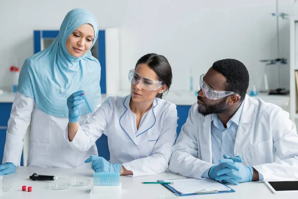 Scientifique tenant une éprouvette près de collègues interraciaux en lunettes de sécurité, pipette électronique et tablette numérique — Photo de stock