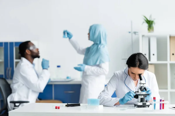 Scientist looking through microscope near blurred interracial colleagues in lab - foto de stock