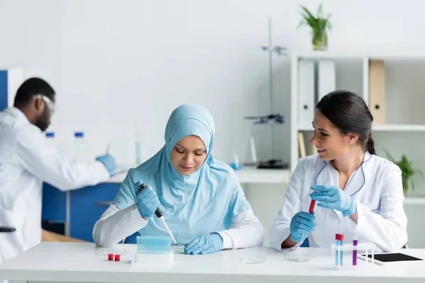 Smiling scientist holding blood sample near arabian colleague with petri dish in laboratory - foto de stock