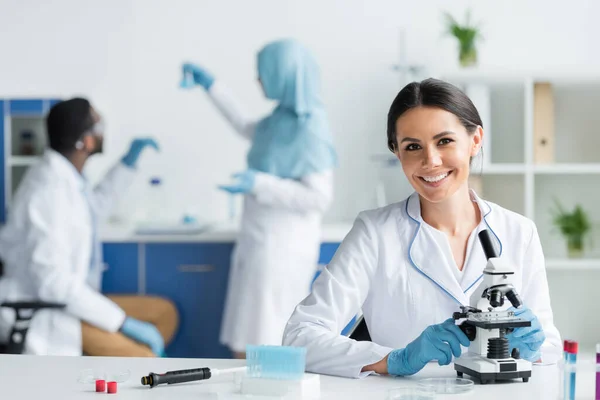 Happy scientist in latex gloves smiling at camera near microscope and test tubes — Stock Photo