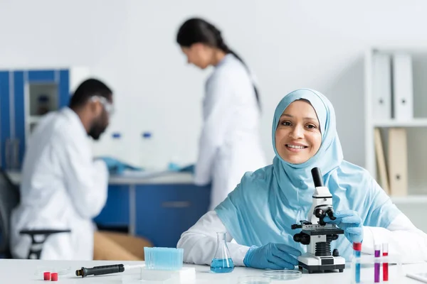 Positive muslim scientist looking at camera near microscope, flask and pipette in lab — Stock Photo