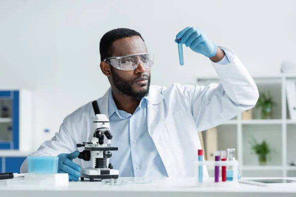 Cientista afro-americano em luvas de látex e óculos de segurança segurando tubo de ensaio perto do microscópio — Fotografia de Stock