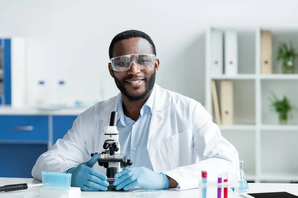 Smiling african american scientist in protective goggles looking at camera near microscope in lab - foto de stock