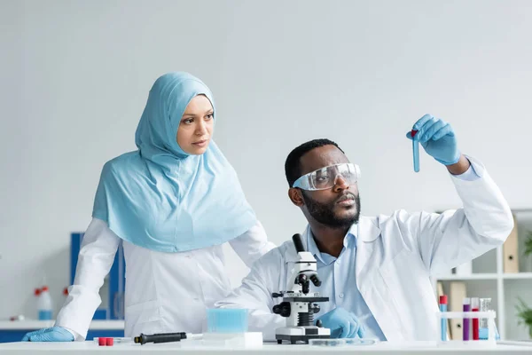 Interracial scientists in latex gloves looking at sample near microscope in laboratory - foto de stock