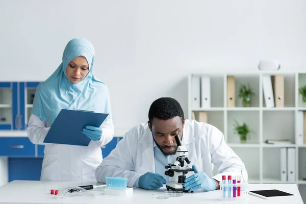 African american scientist looking through microscope near muslim colleague with clipboard - foto de stock