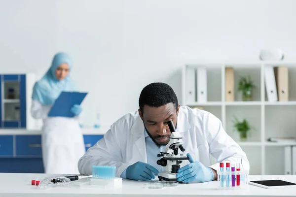 African american scientist in white coat looking through microscope near test tubes and digital tablet - foto de stock