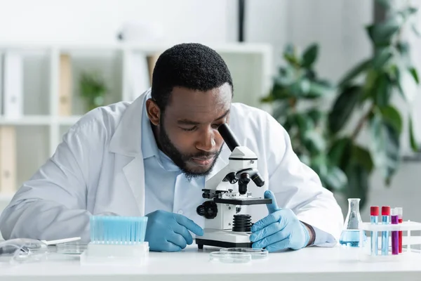Cientista afro-americano em luvas de látex trabalhando com estetoscópio, tubo de ensaio e frasco — Fotografia de Stock