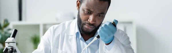 African american scientist in latex glove holding electronic pipette near blurred microscope, banner - foto de stock