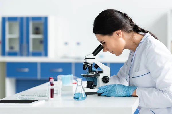 Side view of scientist in latex gloves using microscope near digital tablet with blank screen and equipment — Stock Photo