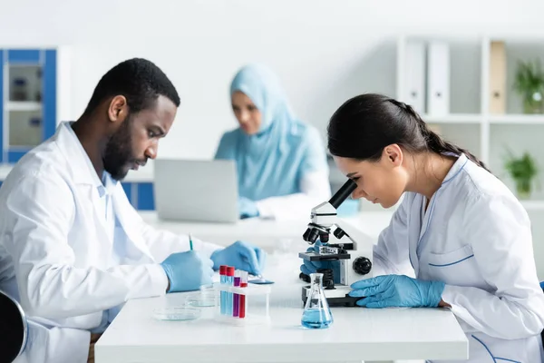 Multiethnic scientists writing on clipboard and working with microscope in lab — Stock Photo