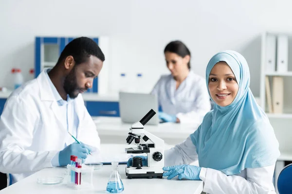 Smiling arabian scientist in hijab looking at camera near microscope and african american colleague in laboratory — Stock Photo