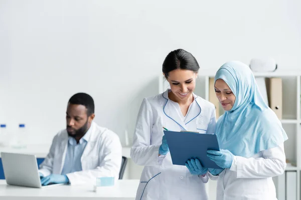Scienziati interrazziali sorridenti mentre tengono appunti vicino a un collega afroamericano offuscato con laptop in laboratorio — Foto stock