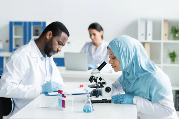 Arabischer Wissenschaftler arbeitet mit Mikroskop in der Nähe von Reagenzgläsern und verschwommenen Kollegen — Stockfoto