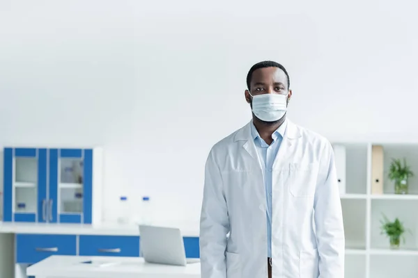 African american scientist in medical mask and white coat looking at camera - foto de stock