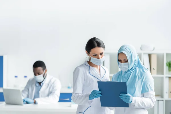 Interracial scientists in medical masks looking at clipboard near african american colleague — Stock Photo