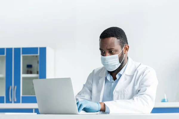 African american scientist in medical mask using laptop in laboratory - foto de stock