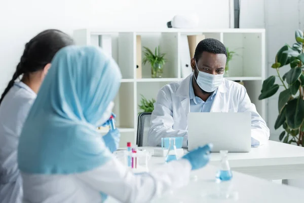 African american scientist in medical mask using laptop near blurred colleagues with equipment — Stock Photo