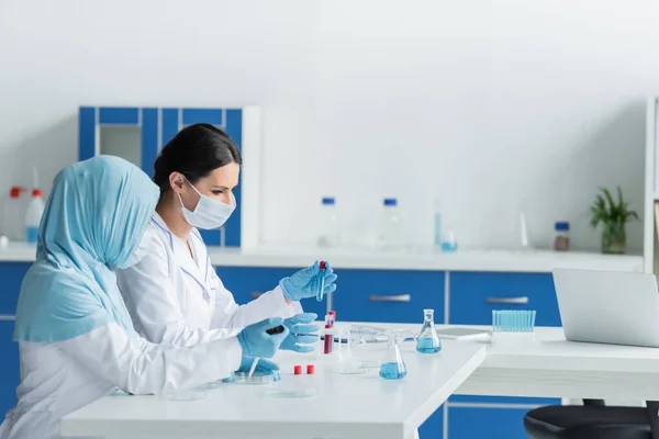 Side view of interracial scientists in protective masks working with medical equipment near laptop in lab - foto de stock