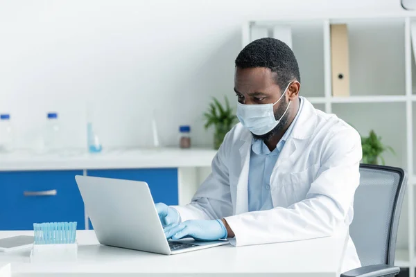 African american scientist in latex gloves and medical mask using laptop near test tubes in lab - foto de stock